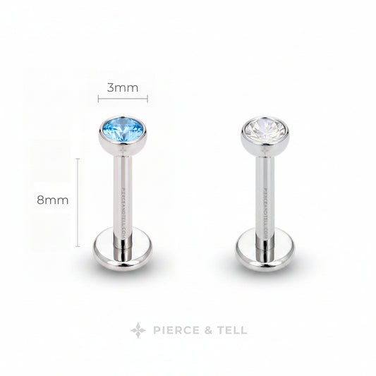 Two silver-toned labret piercing studs on a white background, one with a faceted light blue gemstone and one with a clear gemstone. Dimension labels indicate 3mm head diameter and 8mm post length, with the brand name PIERCE & TELL at the bottom.