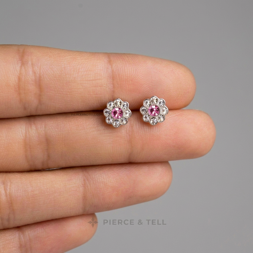 A pair of silver-toned stud earrings with round pink center stones surrounded by small clear stones, shown resting on a person's fingers against a plain gray background. The brand name 'PIERCE & TELL' appears faintly near the bottom.