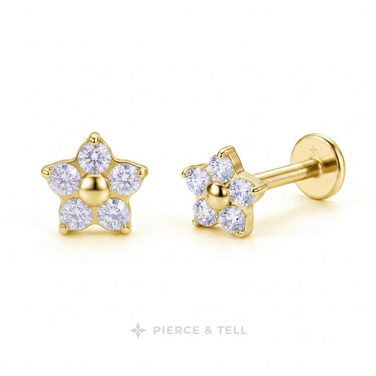 A pair of gold flower-shaped stud earrings with clear round-cut gemstones, shown on a clean white background. One earring is facing forward to show the front face, and the other is turned to show the side profile with the post and backing with pierce & tell logo.