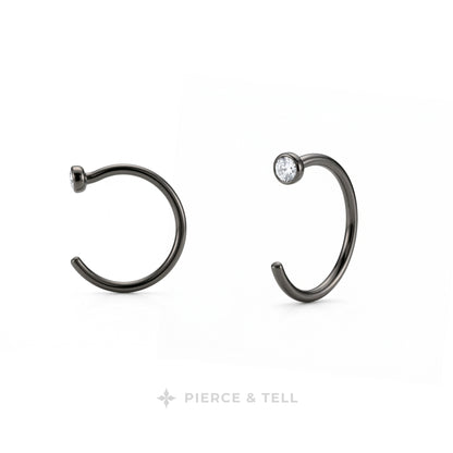 Two dark metallic C-shaped hoop earrings on a white background, one plain and one with a small clear gemstone, with the brand name 'PIERCE & TELL' at the bottom.