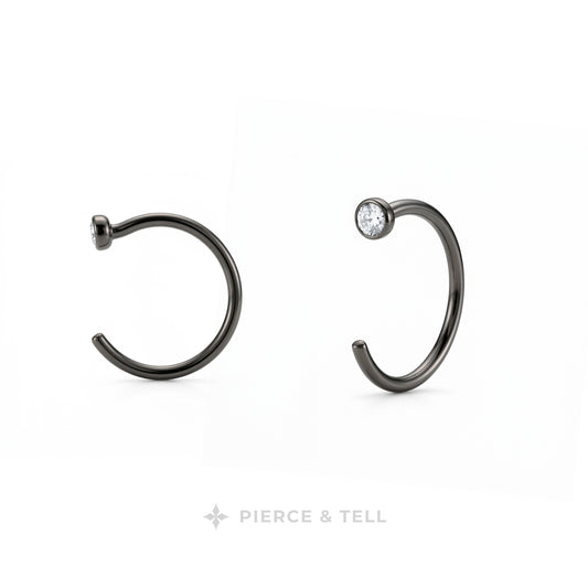 Two dark metallic C-shaped hoop earrings on a white background, one plain and one with a small clear gemstone, with the brand name 'PIERCE & TELL' at the bottom.