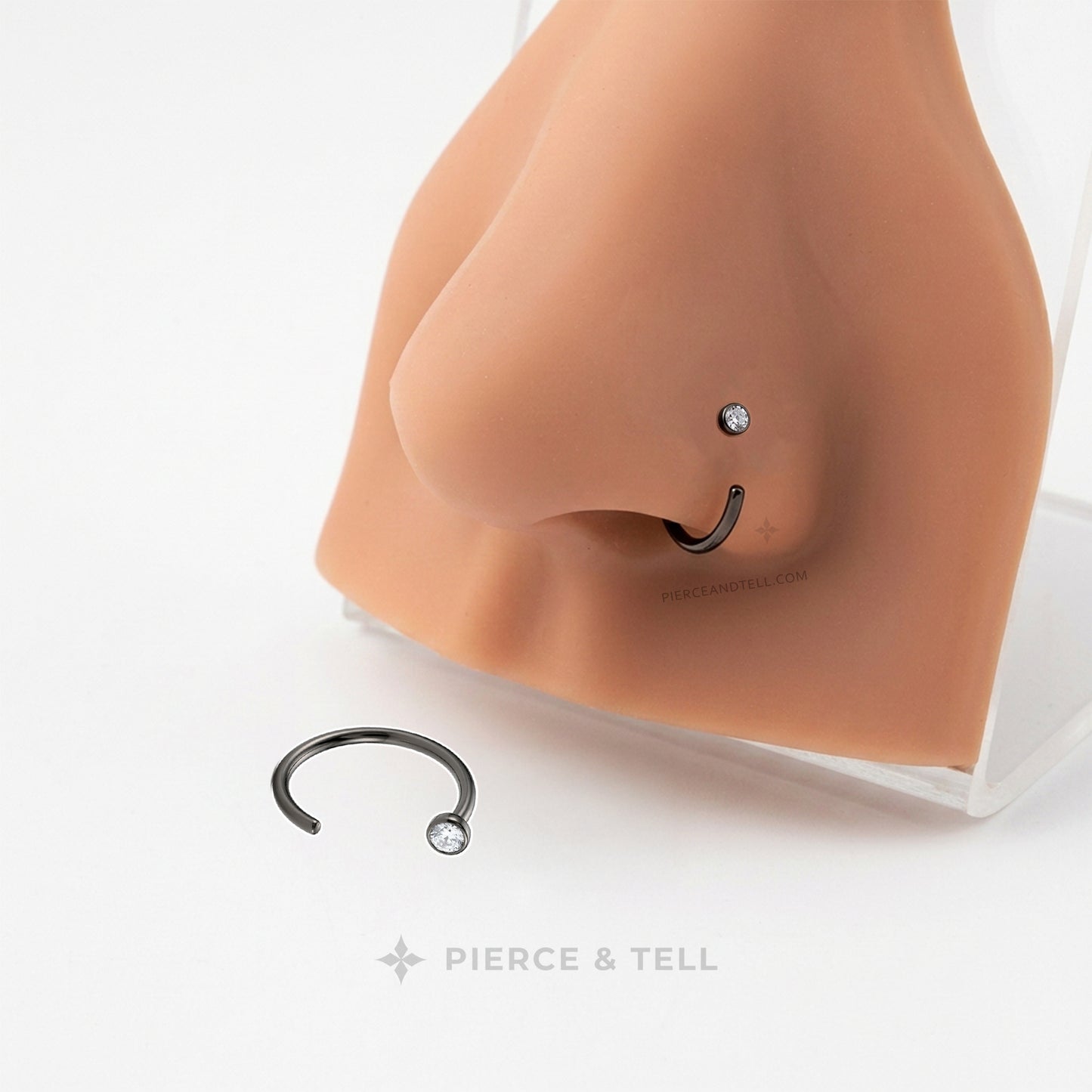 A black metal nose ring with a clear rhinestone stud is displayed on a silicone nose mannequin and also shown loose on a white surface. The brand name 'PIERCE & TELL' is visible at the bottom.