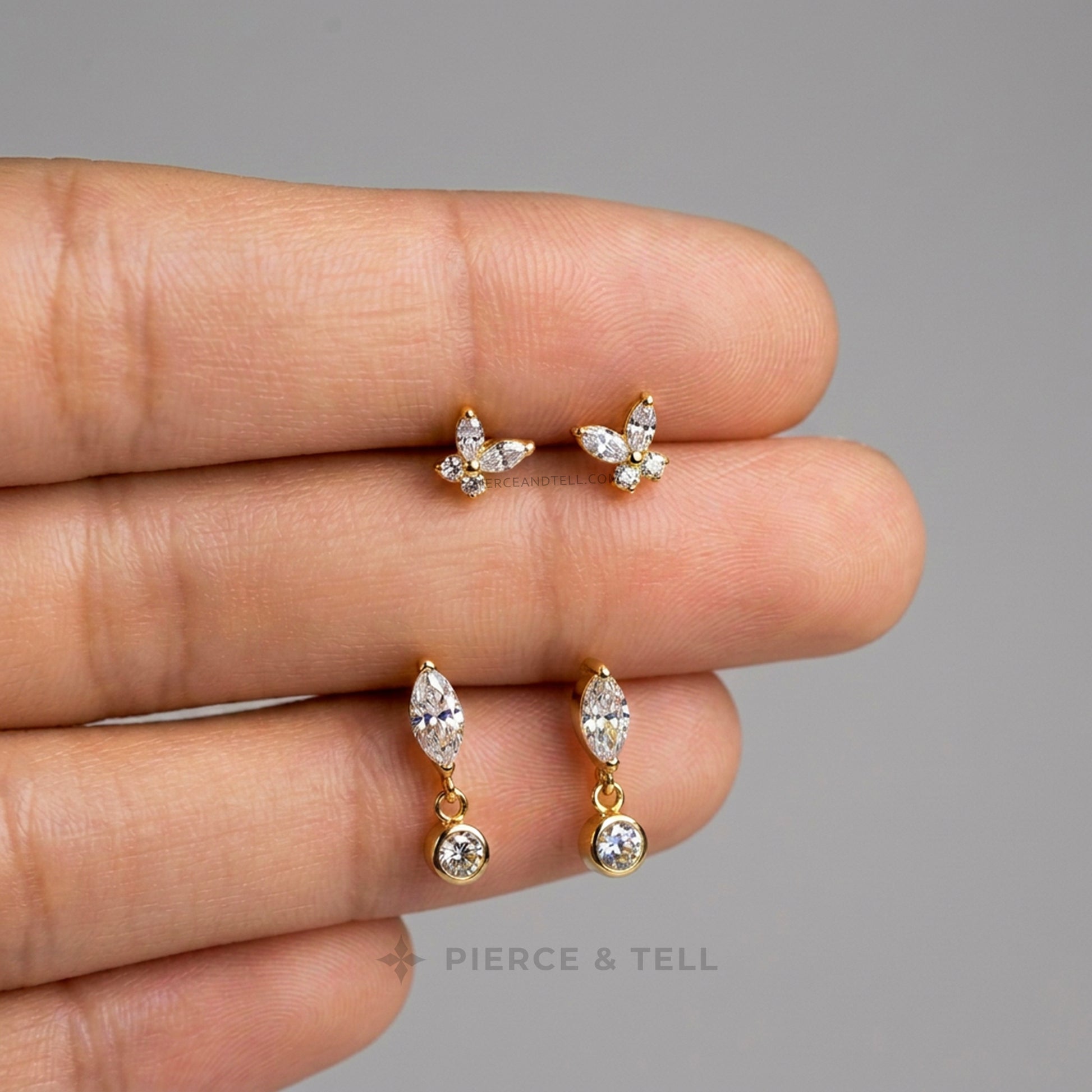 A close-up of a hand holding three pairs of gold-toned earrings against a plain gray background: two small butterfly-shaped stud earrings and two small marquise-shaped dangle earrings with clear stones. The text 'PIERCE & TELL' appears faintly near the bottom.
