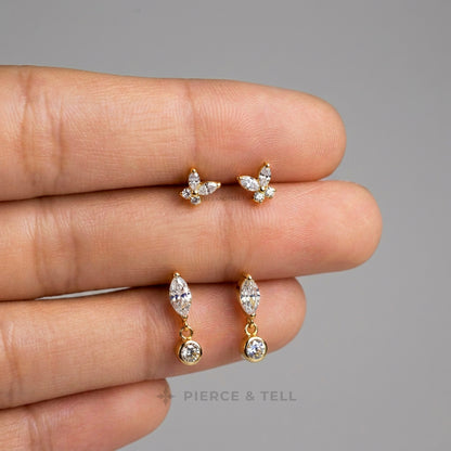 A close-up of a hand holding three pairs of gold-toned earrings against a plain gray background: two small butterfly-shaped stud earrings and two small marquise-shaped dangle earrings with clear stones. The text 'PIERCE & TELL' appears faintly near the bottom.