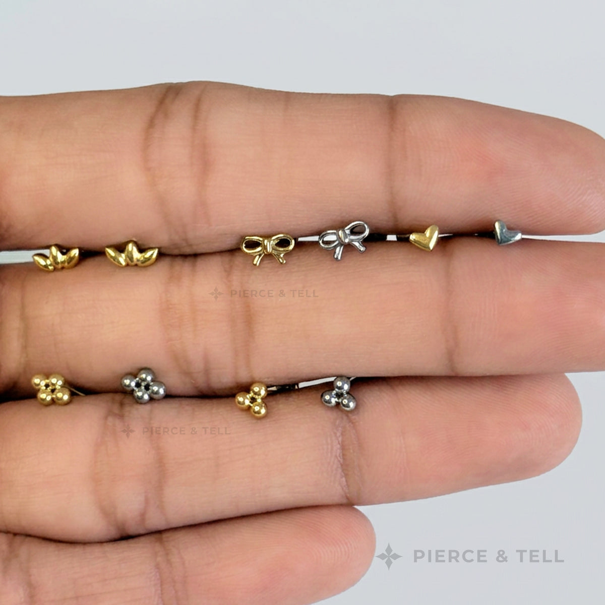 A close-up of a hand displaying two rows of delicate body jewelry. The top row features small gold and silver heart and bow charms on a black cord, while the bottom row shows matching gold and silver flower-shaped charms.