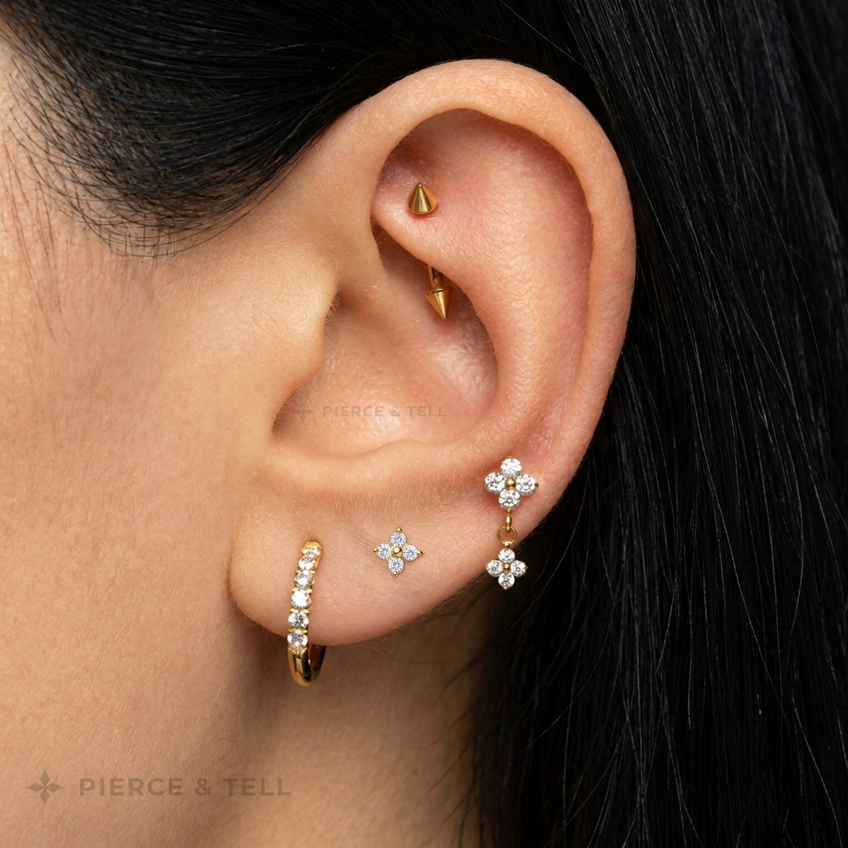 Close-up of a person's ear showcasing multiple gold piercings and earrings, including a conical ear piercing, a vertical huggie hoop with clear stones, a butterfly stud, and a dangling flower earring. The brand name 'Pierce & Tell' is visible as a watermark.