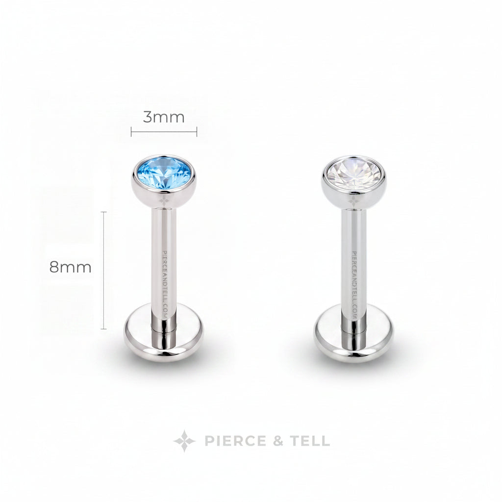 Two silver-toned labret piercing studs on a white background, one with a faceted light blue gemstone and one with a clear gemstone. Dimension labels indicate 3mm head diameter and 8mm post length, with the brand name PIERCE & TELL at the bottom.