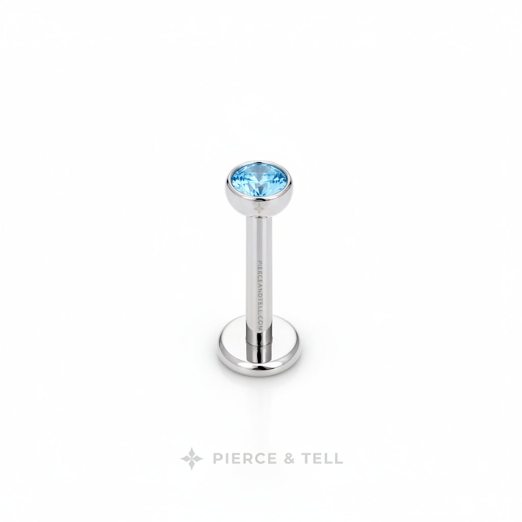 A silver-toned labret piercing barbell with a round bezel-set light blue gemstone, shown on a plain white background with the brand name 'PIERCE & TELL' printed below.