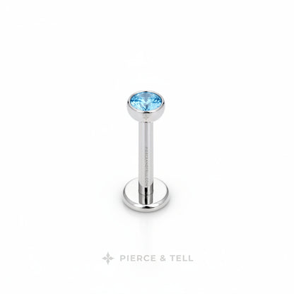 A silver-toned labret piercing barbell with a round bezel-set light blue gemstone, shown on a plain white background with the brand name 'PIERCE & TELL' printed below.