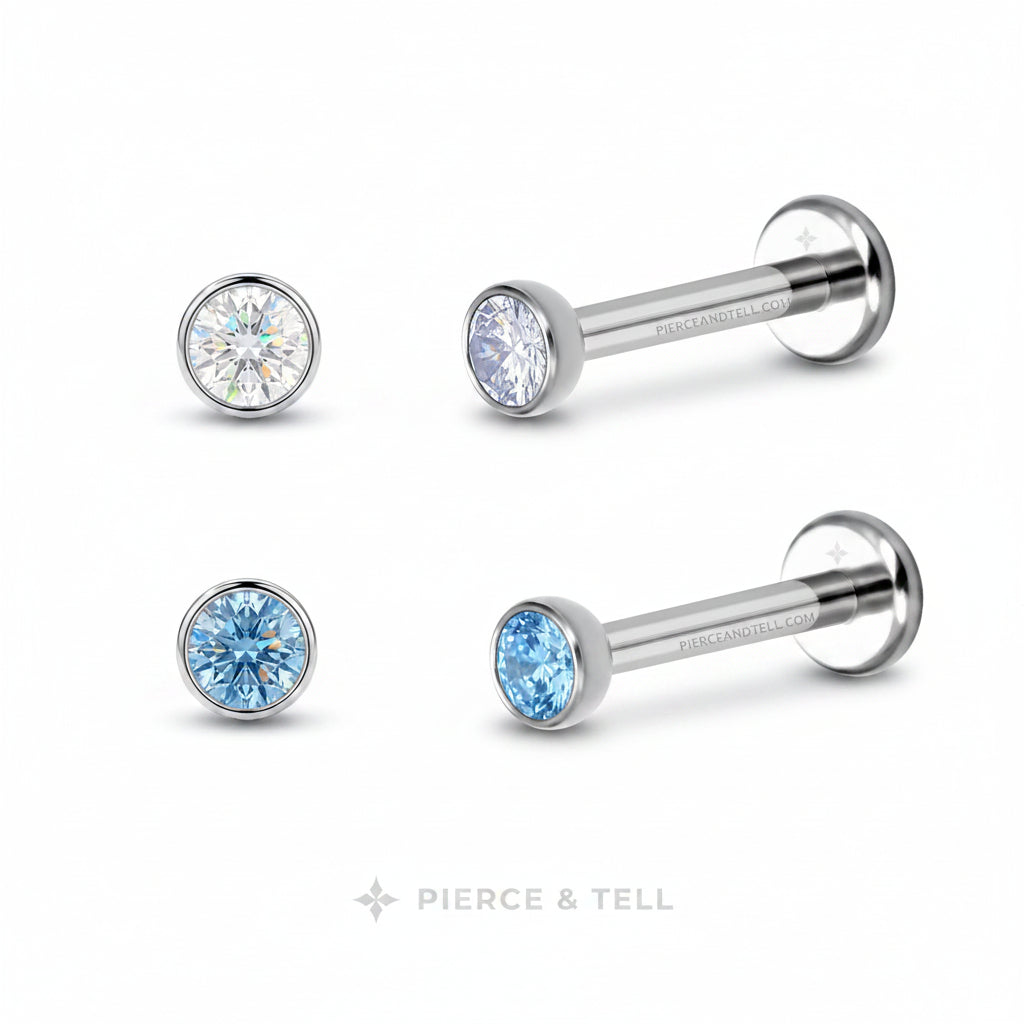 Two silver-toned labret piercing studs from Pierce & Tell, each with a round bezel-set crystal. One stud features a clear faceted crystal, while the other features a light blue faceted crystal.
