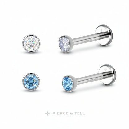 Two silver-toned labret piercing studs from Pierce & Tell, each with a round bezel-set crystal. One stud features a clear faceted crystal, while the other features a light blue faceted crystal.