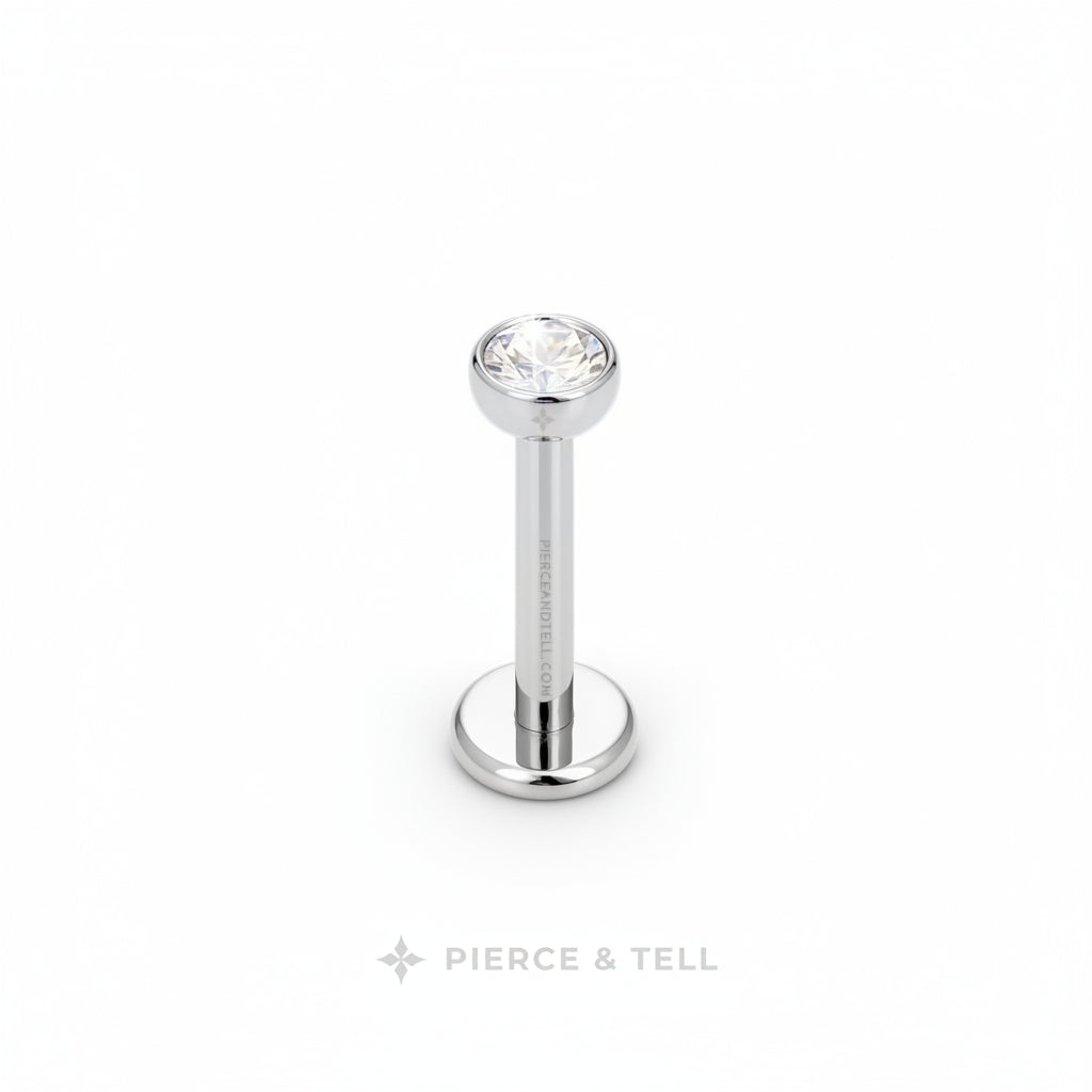 A silver-toned labret piercing stud with a clear faceted gemstone on a white background, featuring the PIERCE & TELL logo below.