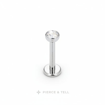 A silver-toned labret piercing stud with a clear faceted gemstone on a white background, featuring the PIERCE & TELL logo below.