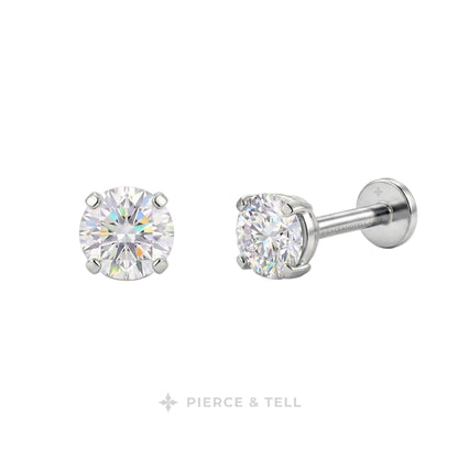 A pair of round-cut diamond stud earrings in a silver-toned setting, with one earring facing forward and the other angled to show the post and backing. The brand name 'PIERCE & TELL' is printed below the earrings on a plain white background.
