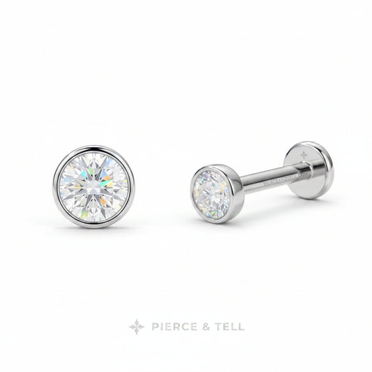 A pair of silver-toned labret stud earrings with round bezel-set clear gemstones, shown from front and side angles on a white background with the PIERCE & TELL logo at the bottom.